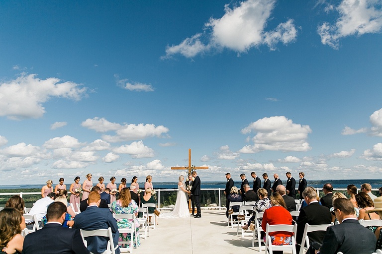 A wide photograph of a wedding ceremony on a sunny day with large white clouds and Lake Michigan in the background