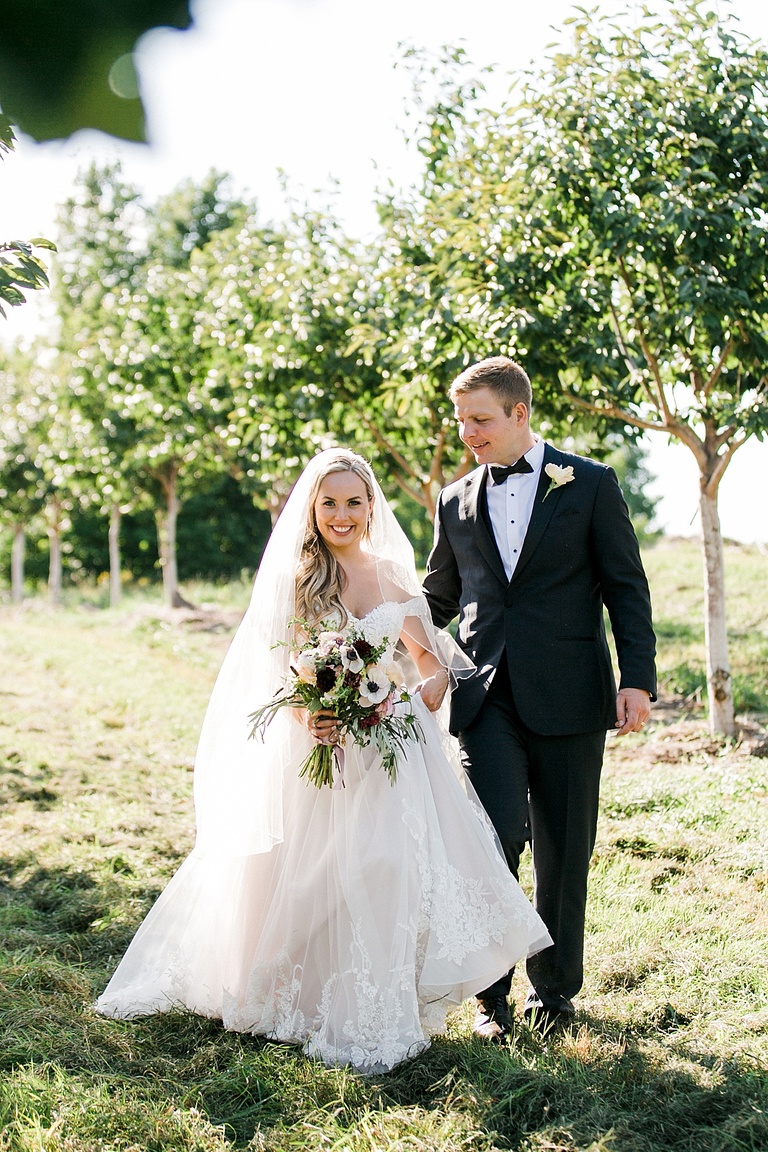 A bride and groom walking through a grassy orchard