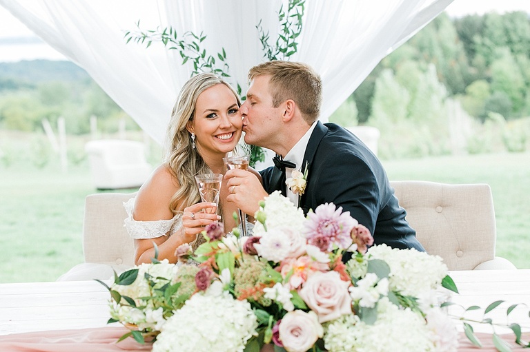 A groom kissing his bride at their wedding reception while holding change at the sweetheart table surrounded by a flower arrangment