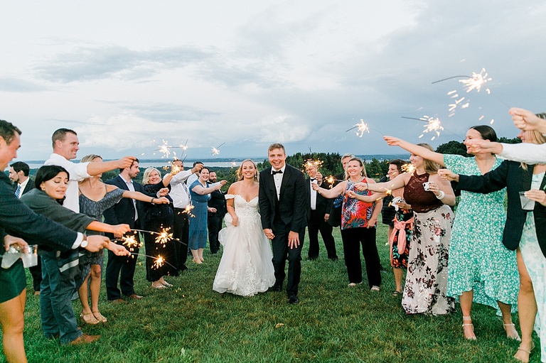 A bride and groom running through sparklers that their guests are holding at dusk