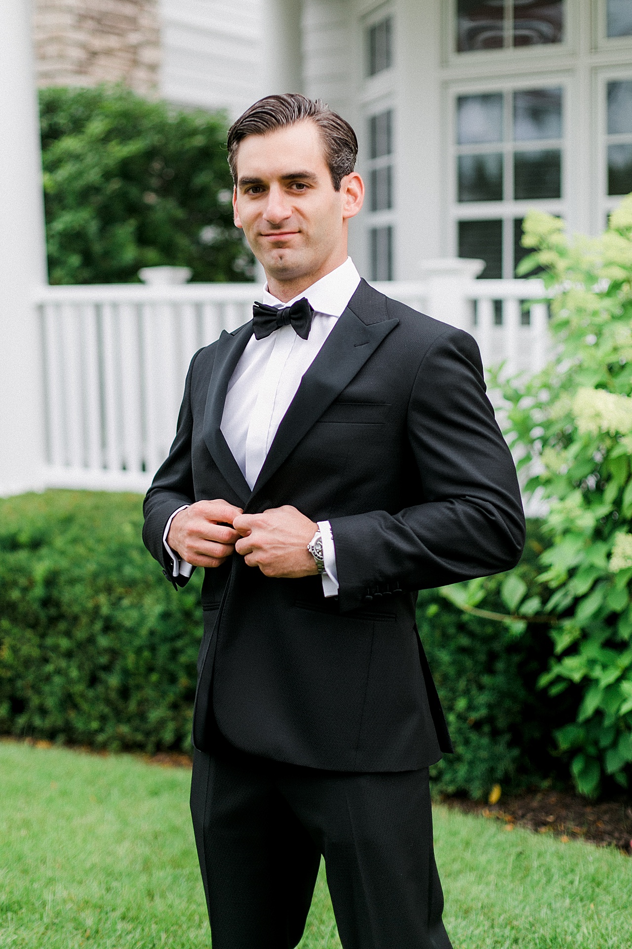 Portrait of a groom buttoning his jacket with white fences and greenery in the background