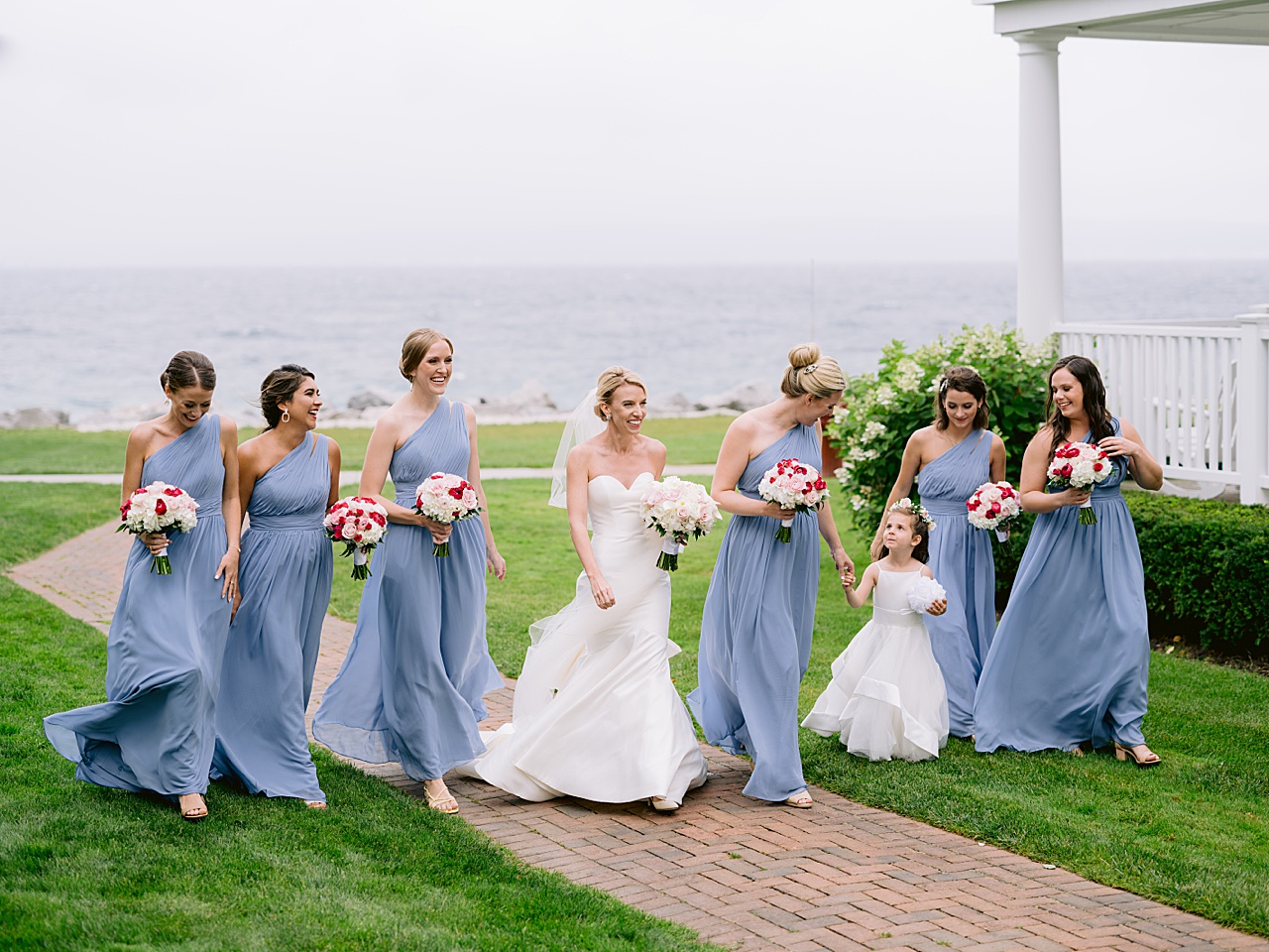 A bride and bridesmaids in blue walk in the wind next to Lake Michigan