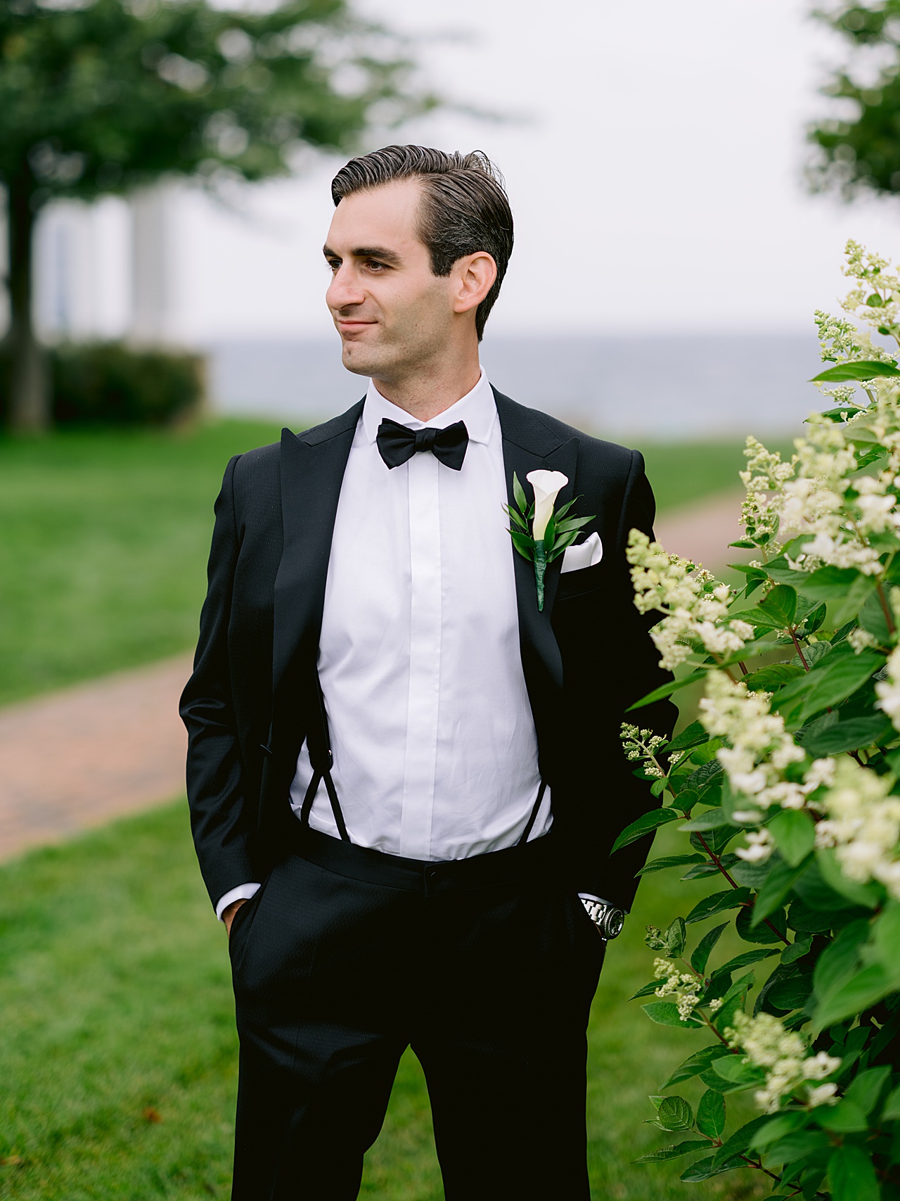 A groom stands with hands in pockets looking of in the distance while surrounded by greenery