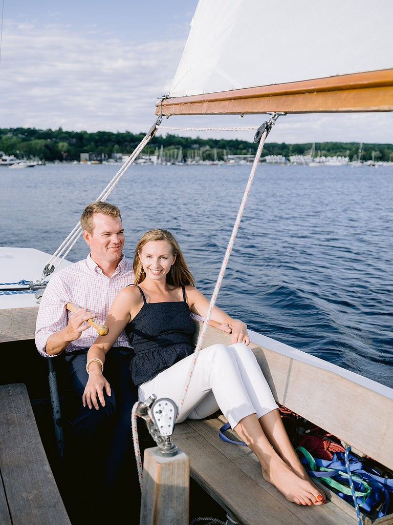 A woman in a navy top and white pants leaning agains her fiancé in a sailboat