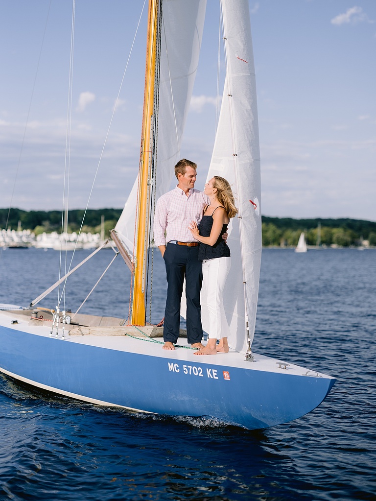 An engaged couple standing on the front of a sailboat on a lake