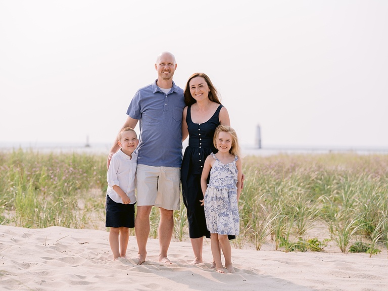 A family stands smiling in the sand and sunshine with Lake Michigan behind them