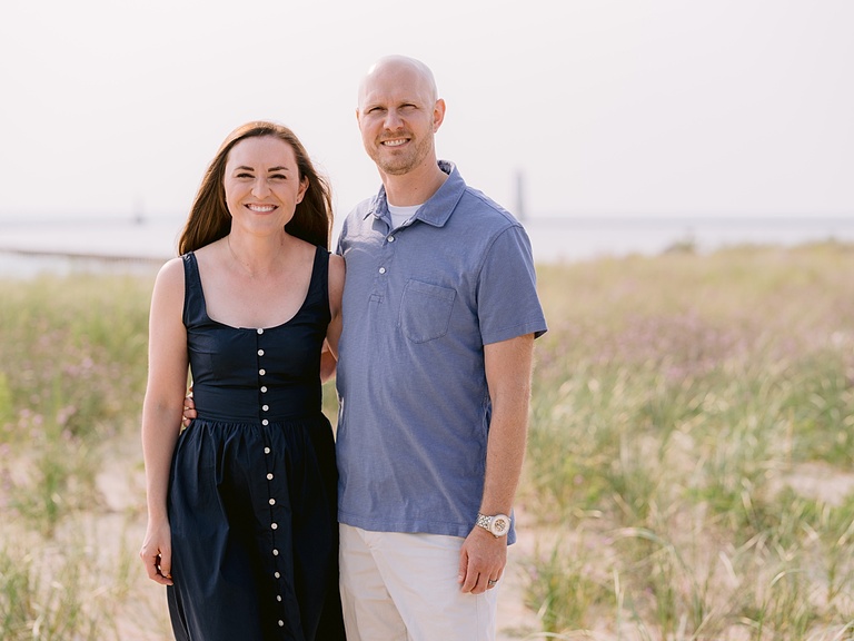 A man and woman smile with an arm around each other with tall grass in the background