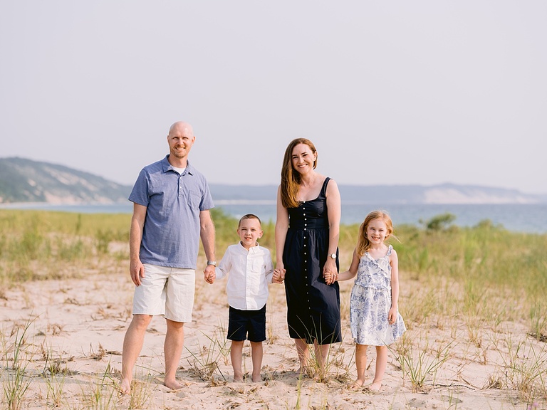 A family of four standing together while holding hands with Lake Michigan and dunes in the background