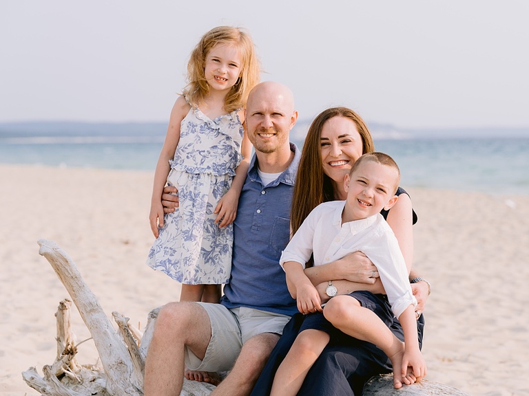 A family of four smiles while sitting on driftwood on a sandy beach near Traverse City