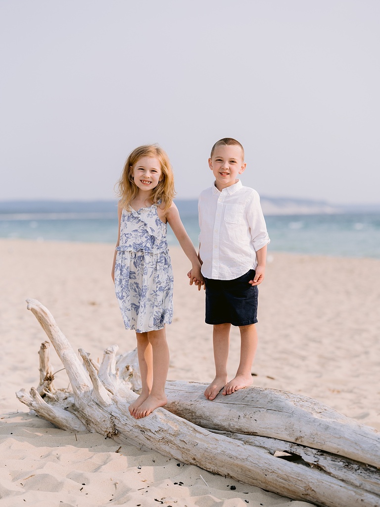 A girl and boy stand on a piece of driftwood and smile on a gorgeous beach in Northern Michigan