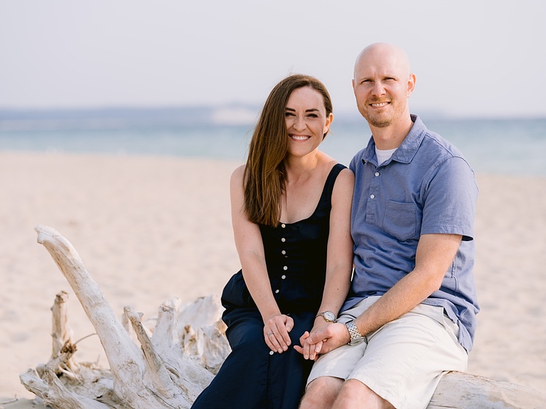 A husband and wife sitting on a piece of driftwood while holding hands at a Michigan beach