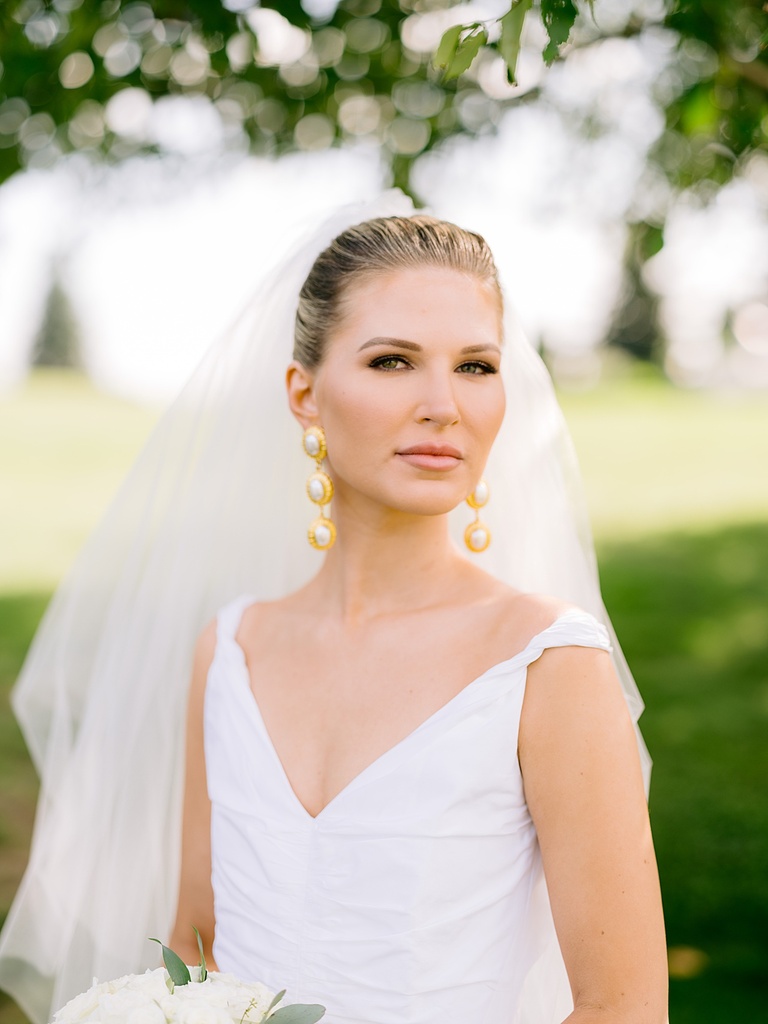 A bride looking at the camera in her white wedding dress, gold Chanel earrings, and long white veil