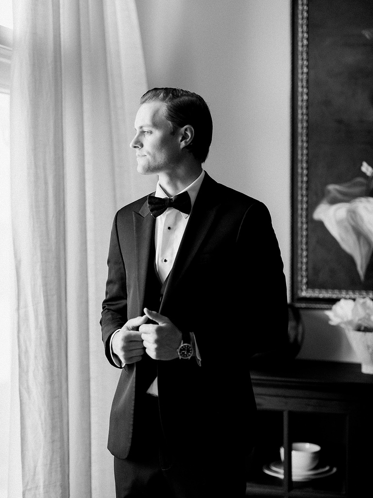 A groom looking out of a widow while putting on his tux jacket from Men's Wearhouse