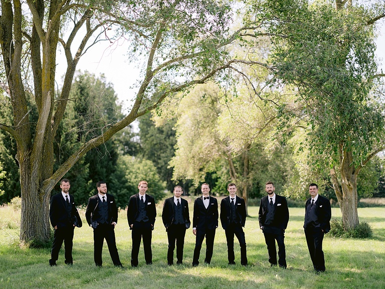 A groom taking portraits in a sunny, grassy field with his groomsmen in Ubly, Michigan