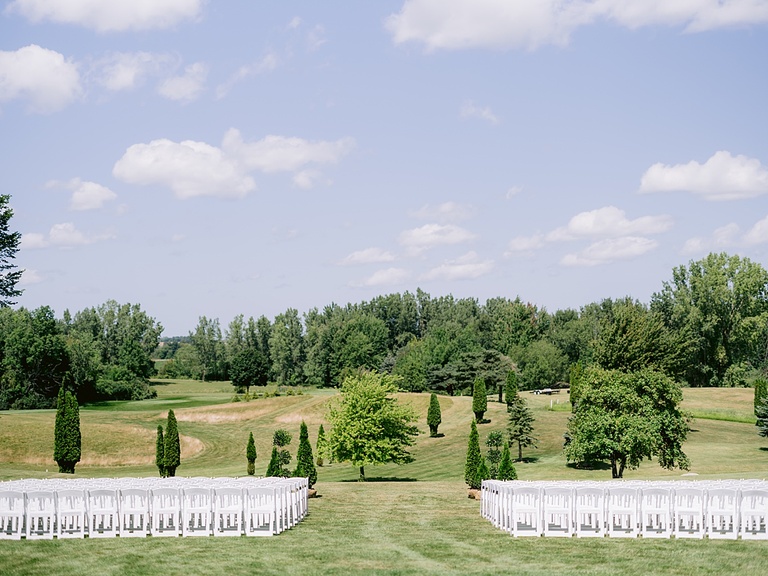 Ceremony location in a grassy filed with trees, blue skies, and white chairs