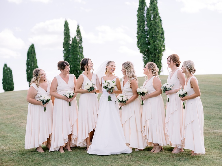 A bride laughing and smiling with her bridesmaids in a open field with tall trees