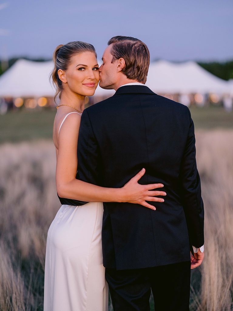 A bride looking at the camera while her groom kisses her cheek with the wedding reception tent in the background