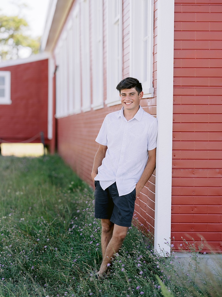 A high school senior boy leans against a red building and smiles with his hands in his pockets