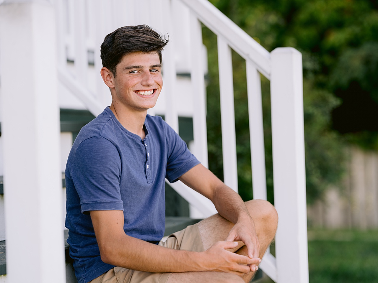 A boy in a blue shirt smiles while sitting on steps in Glen Arbor for senior portraits