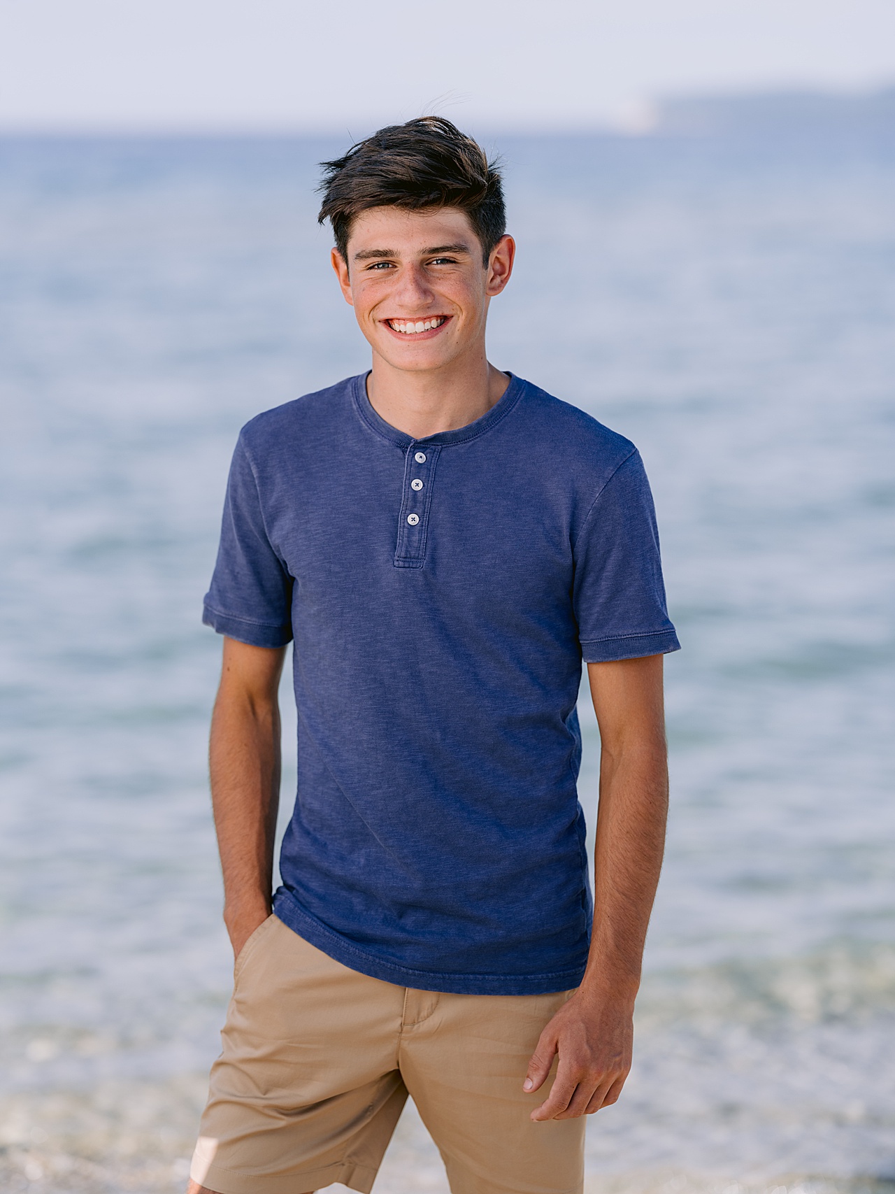 A boy stands with one hand in his pocket with a clear Michigan lake behind him in summer
