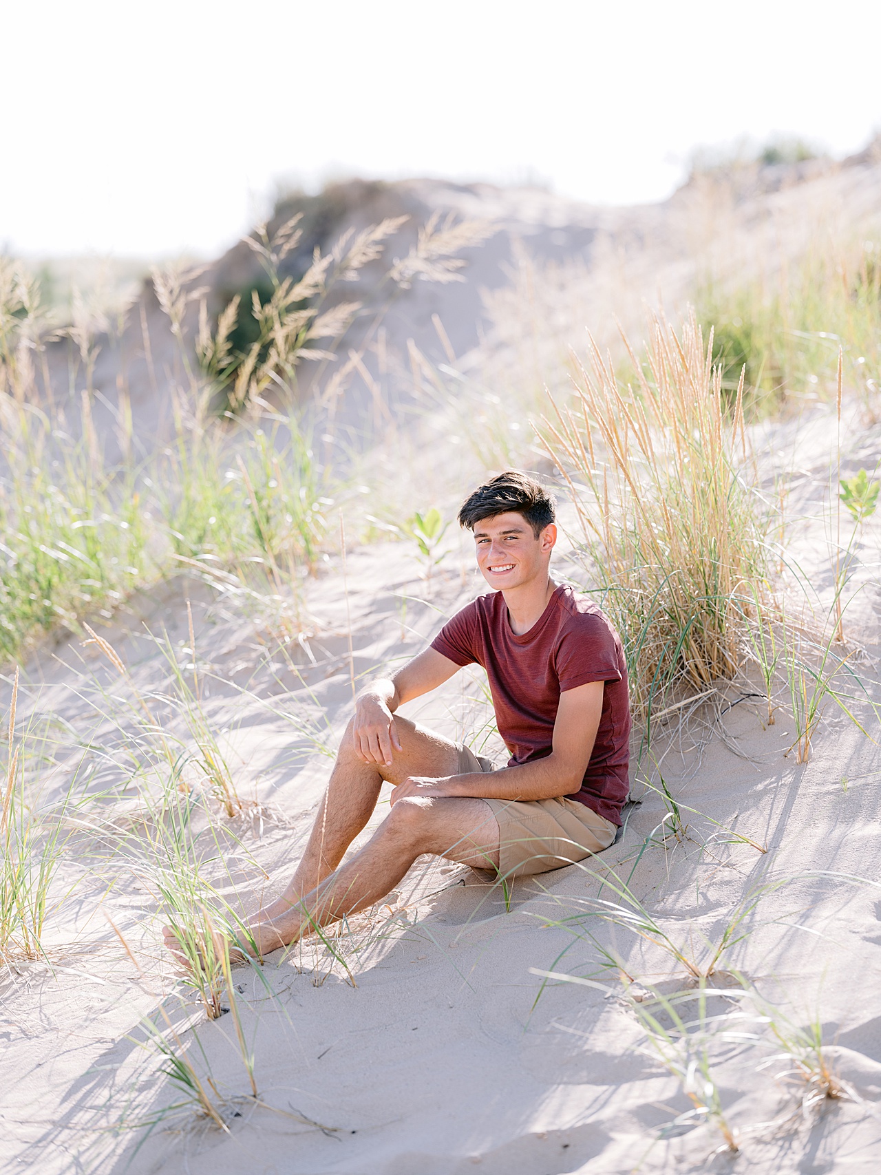 A boy sits in the white sand of a beach and smiles for senior portraits in Glen Arbor