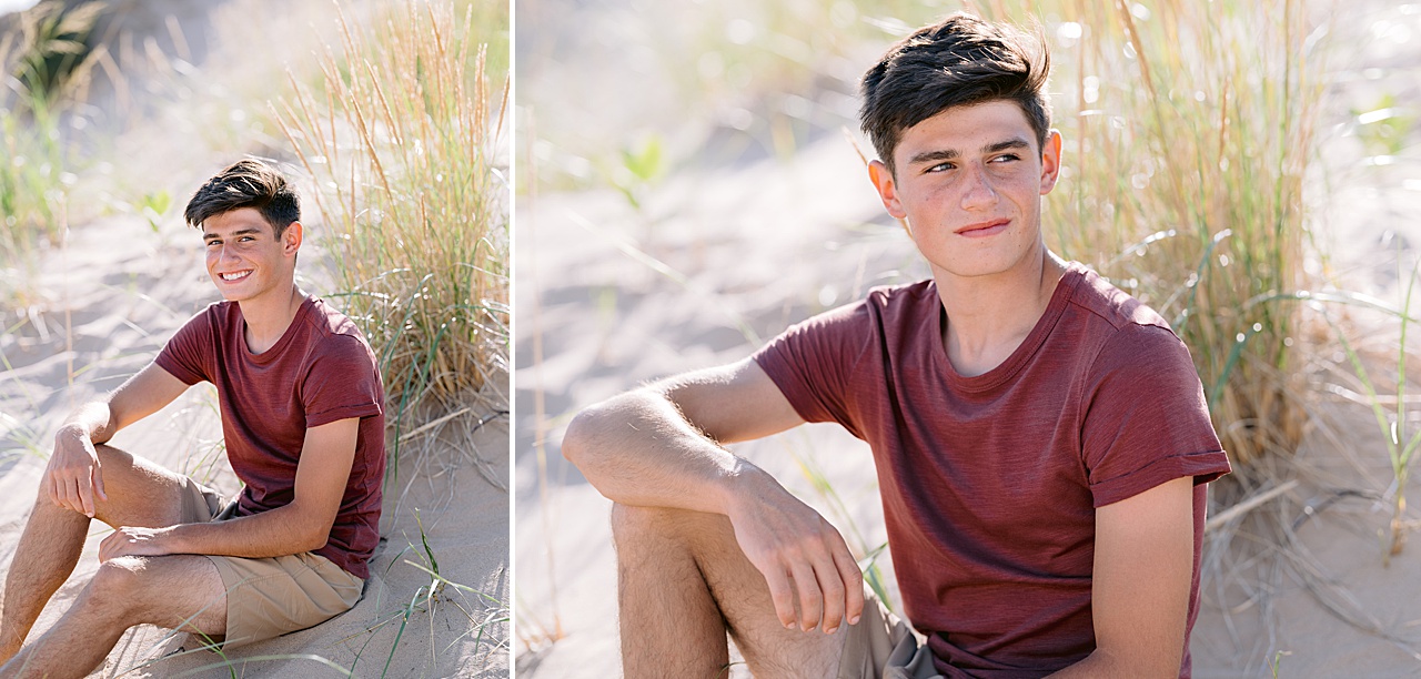 A young man poses for senior portraits while sitting in sand and tall grass