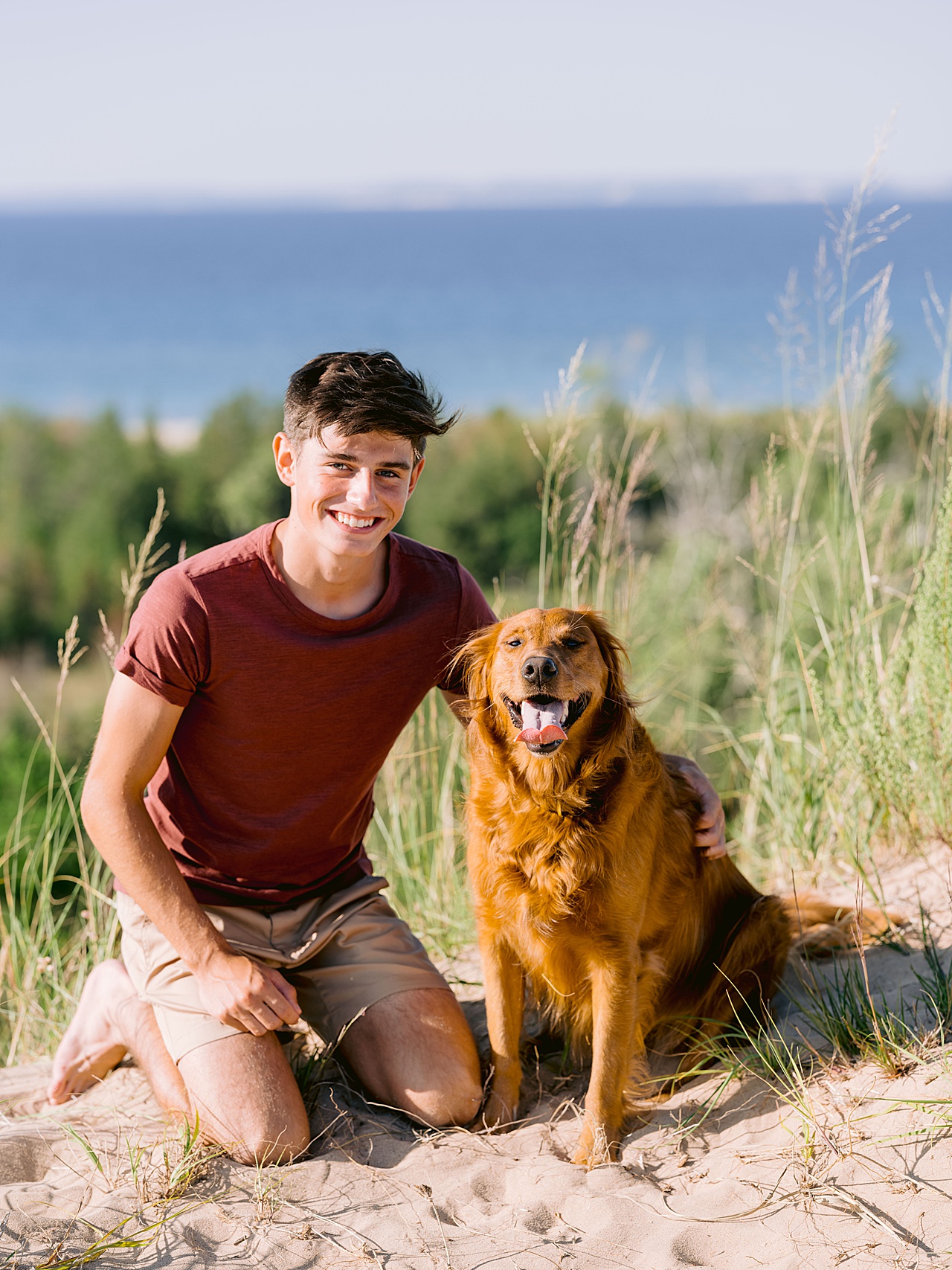 A boy sits on the sand in a red shirt and smiles with his dog for senior portraits