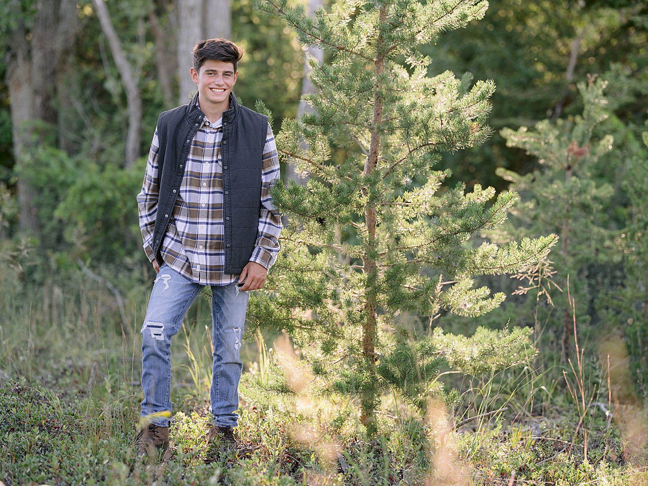 A high school senior poses for photos while standing beside a young pine tree in Michigan