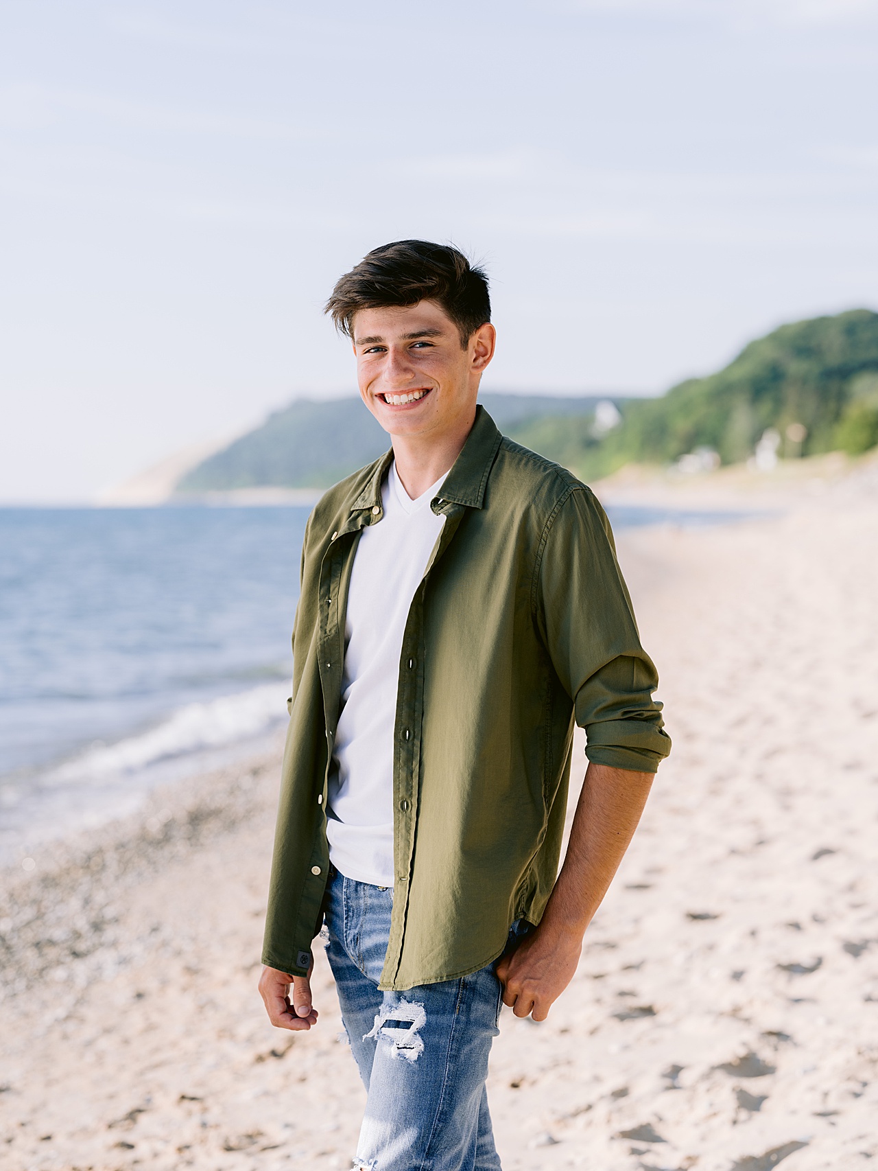 A brunette boy stands smiling next to Lake Michigan for senior pictures