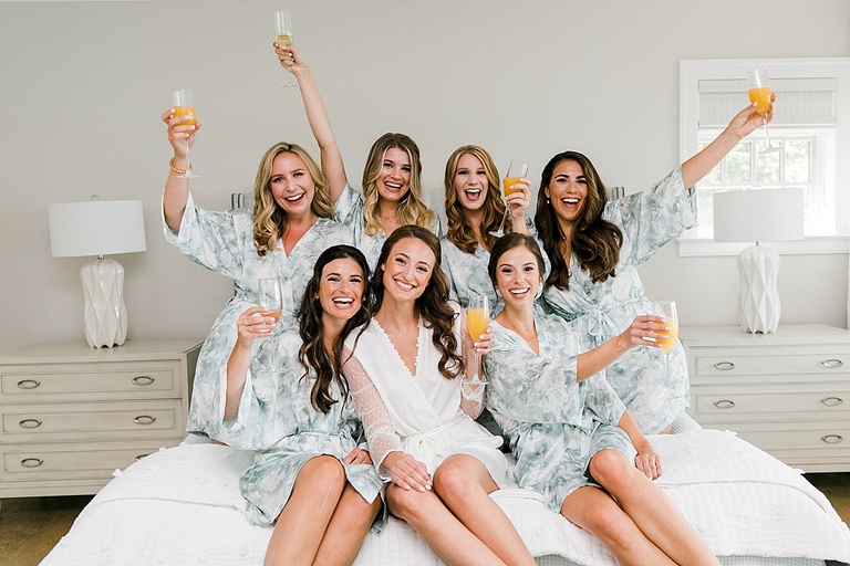 A bride and her bridesmaids holding mimosas together on a bed and clinking glasses