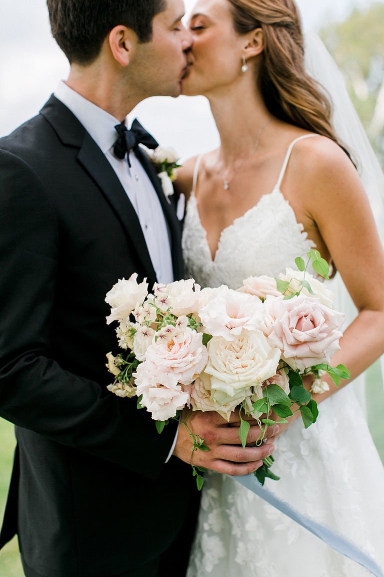 A bride and groom kissing while holding a bridal bouquet with white and light pink flowers