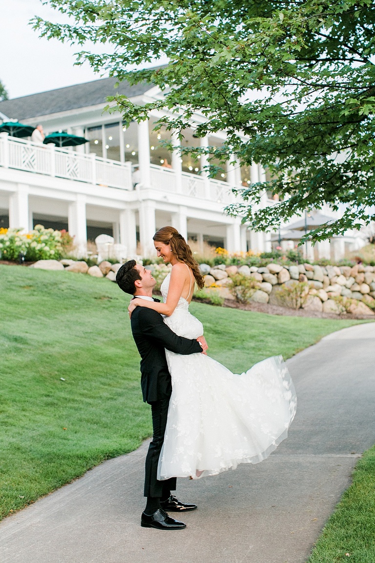 A groom picking up his bride and smiling at her with the wedding venue in the background