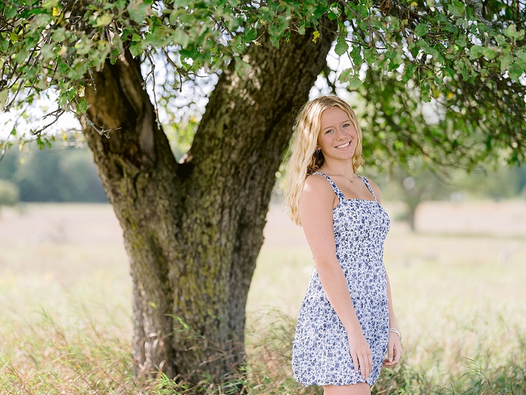 A girl stands smiling for senior portraits under a tree on a sunny summer day