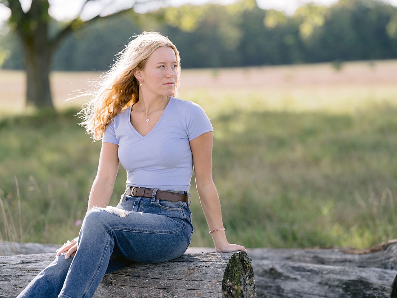 A girl is sitting on a log beside a green field on a summer day in Oneida
