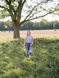 A high school senior girl poses in the tall grass in front of a beautiful tree