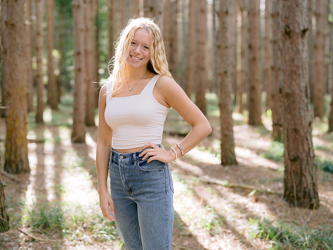 A girl stands smiling with tall pine trees standing behind her
