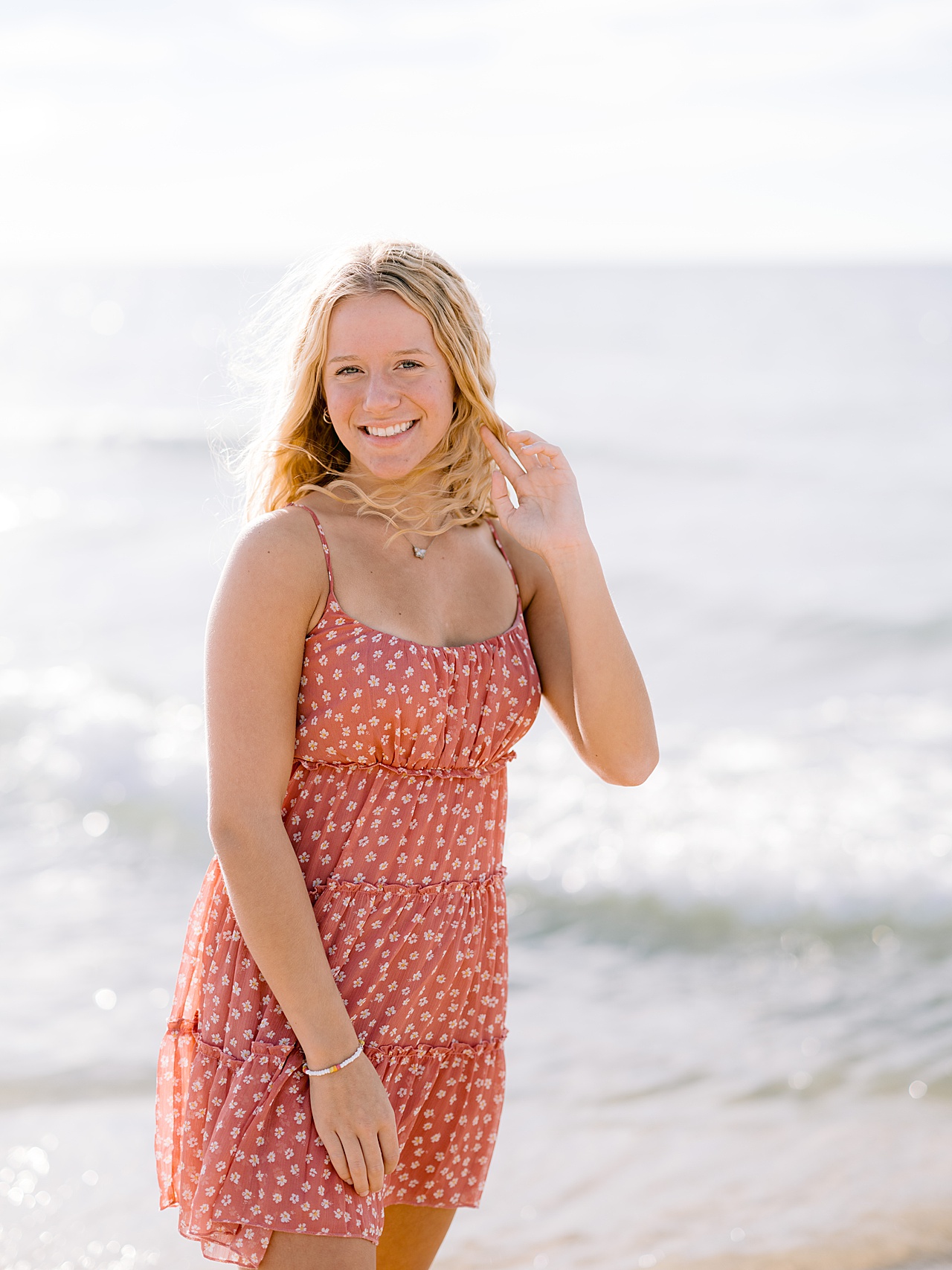 A girl stands smiling while the wind blows her hair with the water of a lake behind her