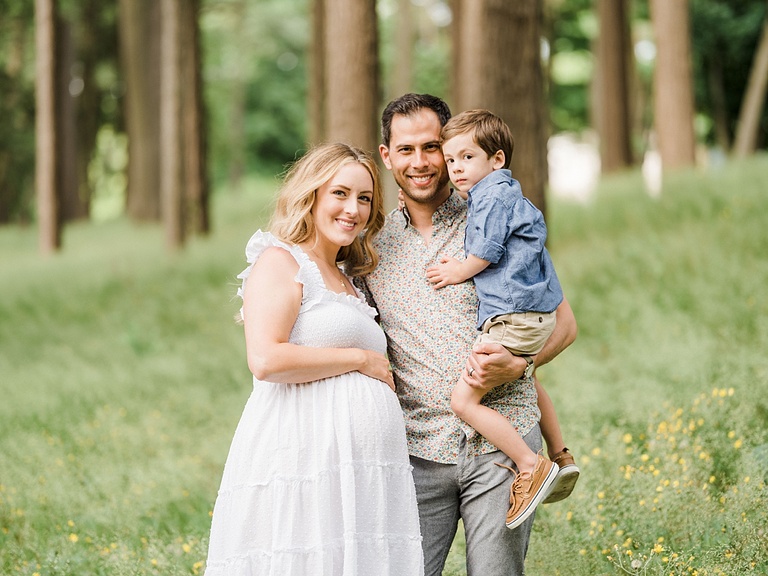 A man and woman smile while holding their toddler boy in a forest in Michigan