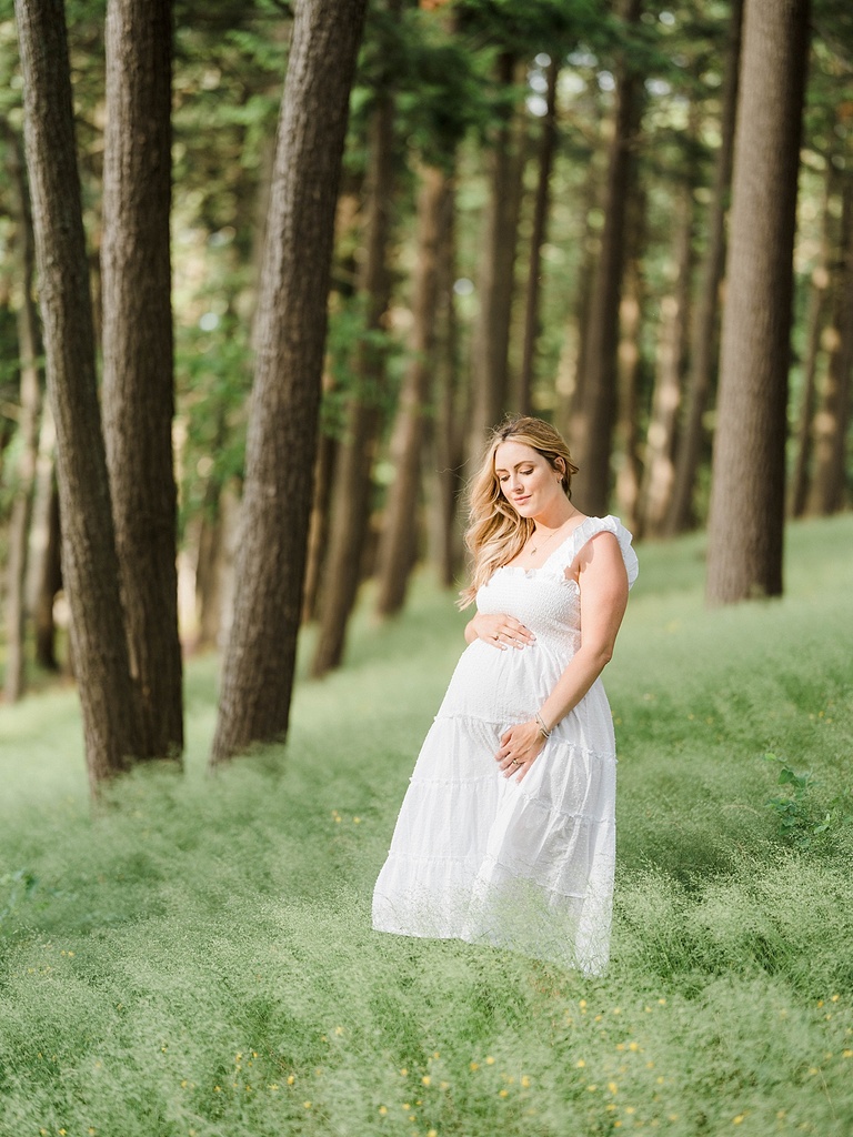 A pregnant woman in a white dress holds her belly while standing in a field of flowers