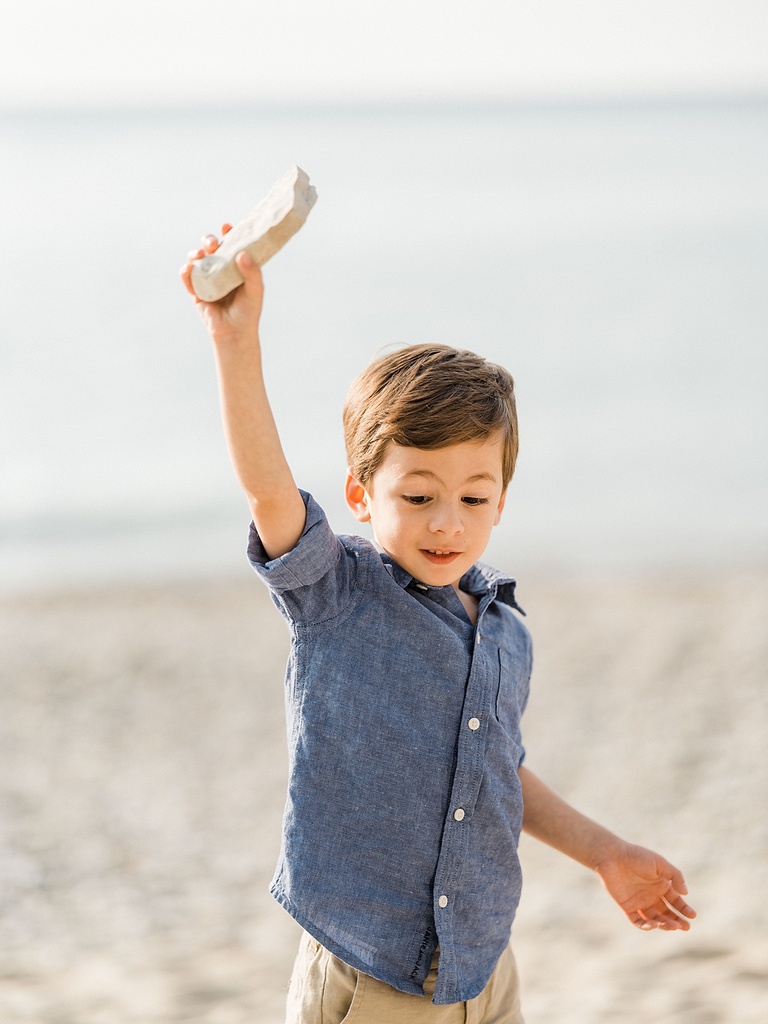 A toddler boy raises a rock over his head to throw in on a sandy beach in northern michigan