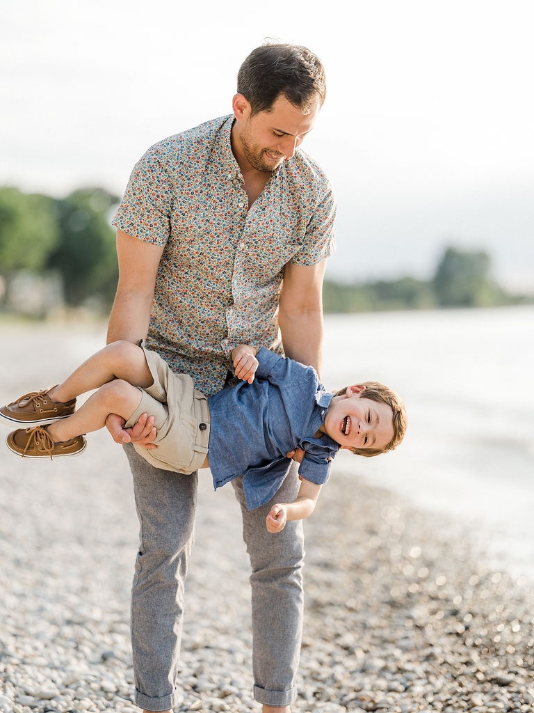 A father curls his son while the young boy laughs on a rocky beach in Charlevoix