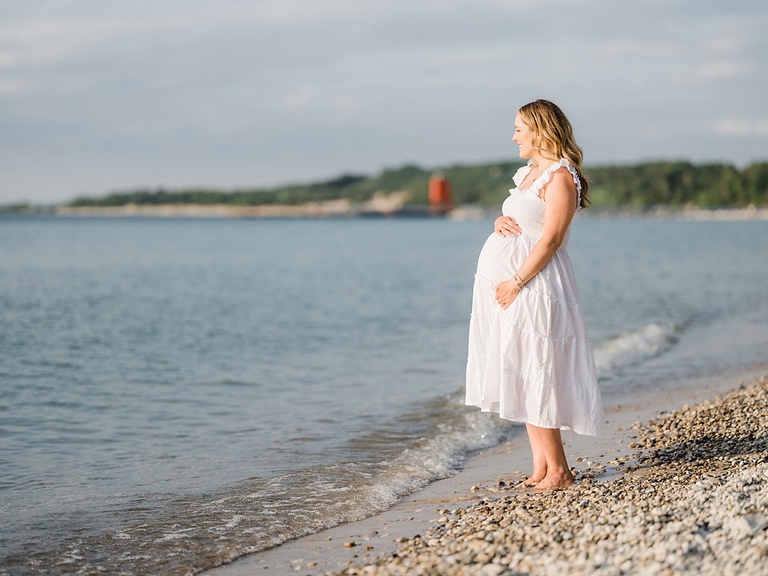 A pregnant mother in a white gown stand on a rocky beach and looks out over Lake Charlevoix