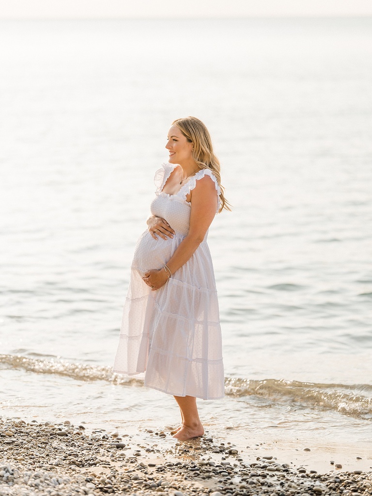 A woman dressed in white stands smiling on a rocky beach in Michigan in the sunshine