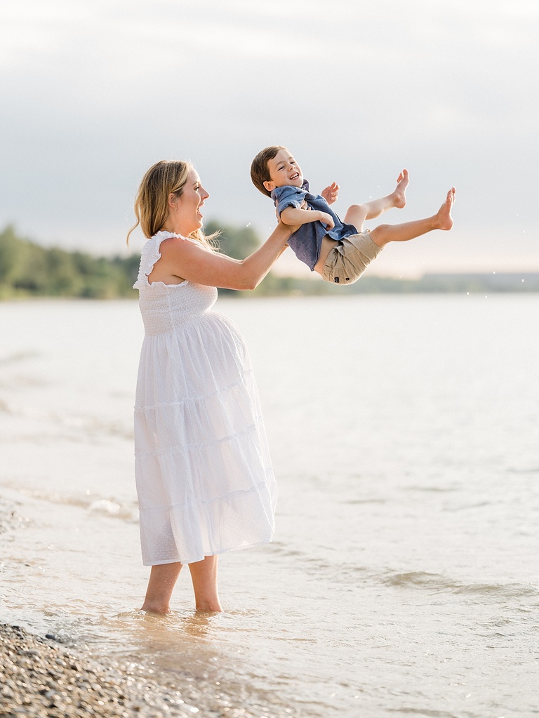 An expectant mother and son smile and laugh while she swings him into the air on Walloon Lake