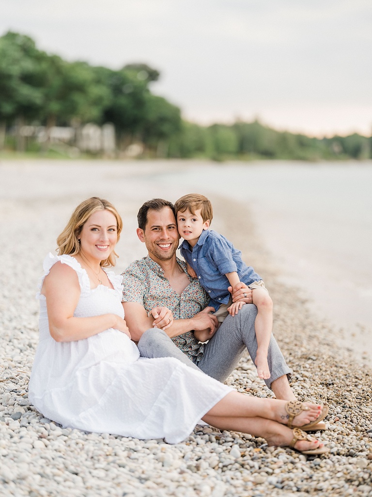 A family of three sits together smiling on a rocky beach beside Lake Walloon