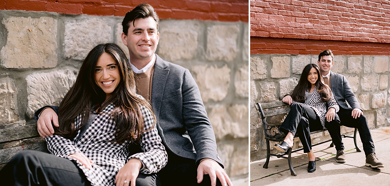 An engaged couple pose on an iron bench in front of a stone and brick building