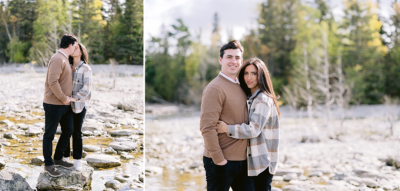 A couple posing for engagement portraits on a rocky shore with a forest behind them