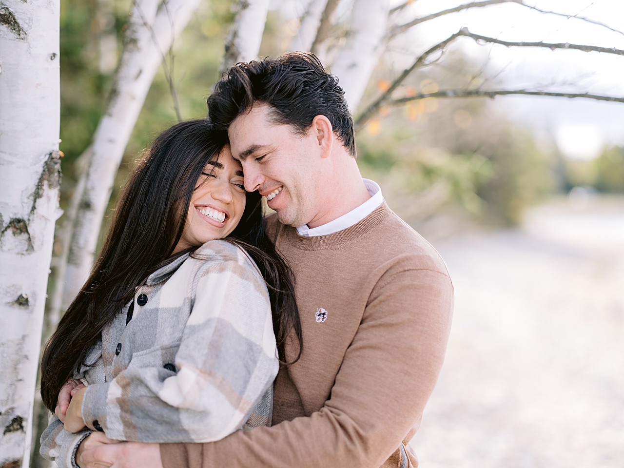 An engaged couple smile and snuggle near a birch tree in Petoskey
