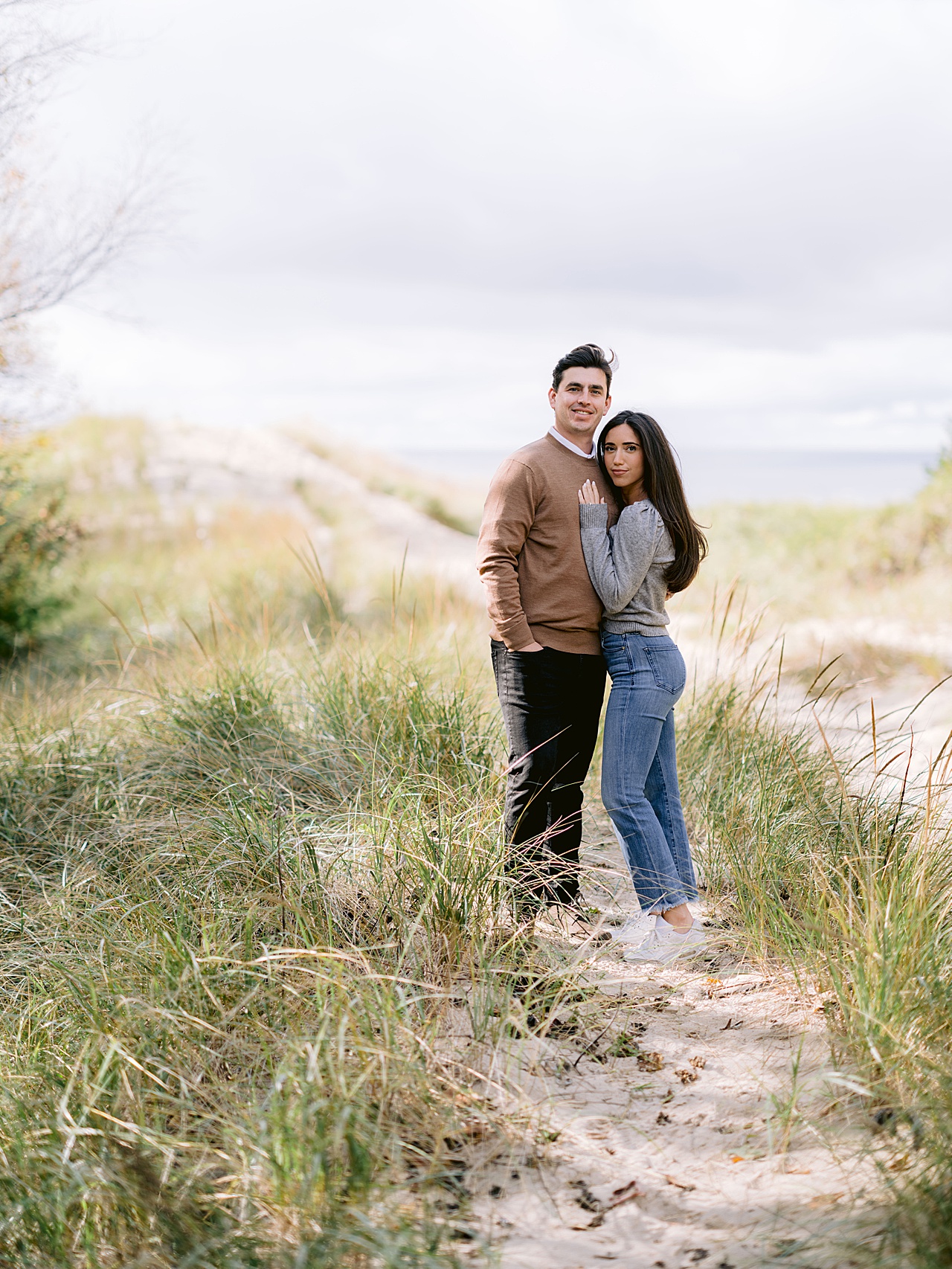A couple pose for engagement portraits with a sandy hill behind them near Lake Michigan
