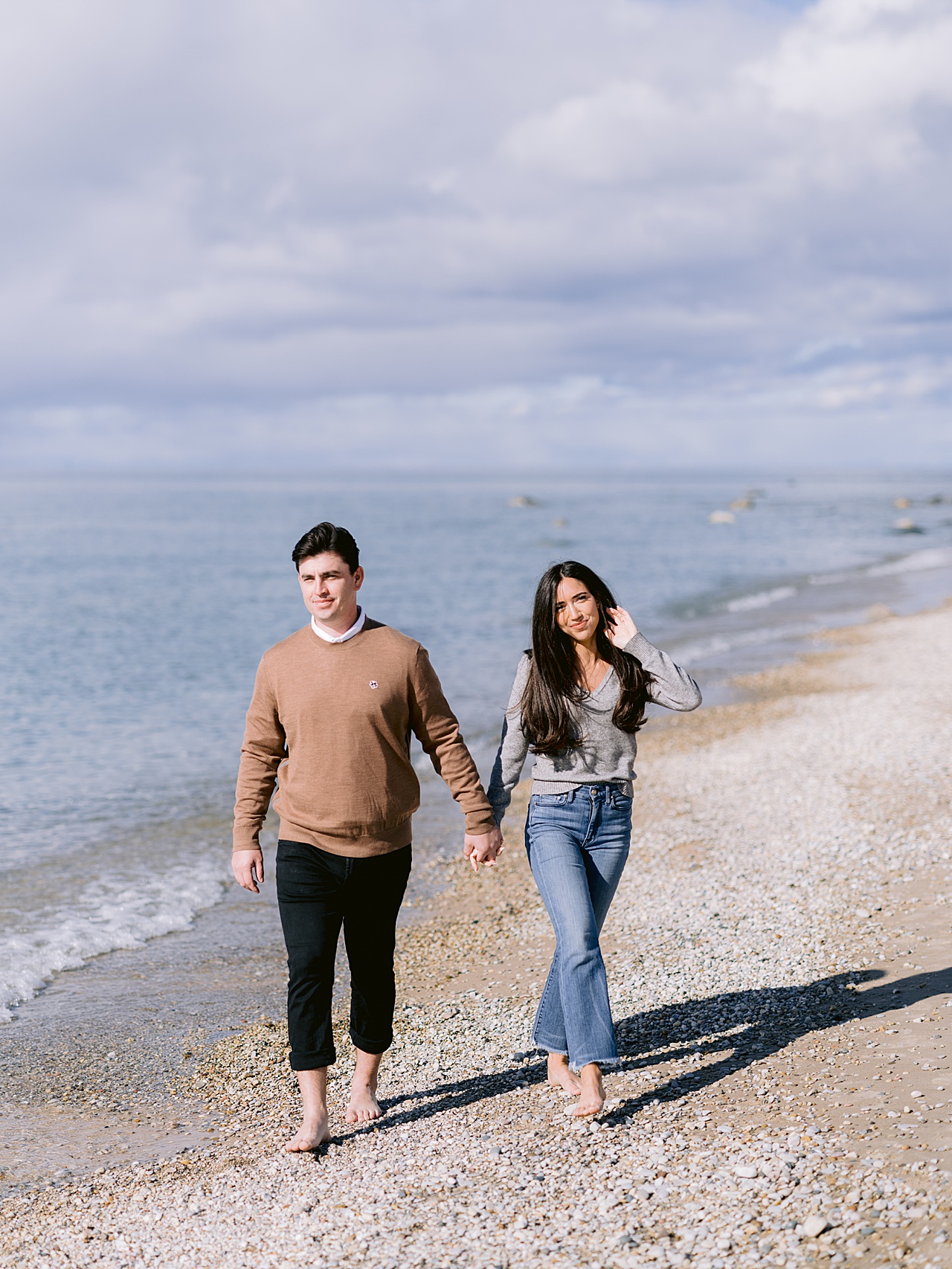 A couple walks a rocky beach barefoot while holding hands